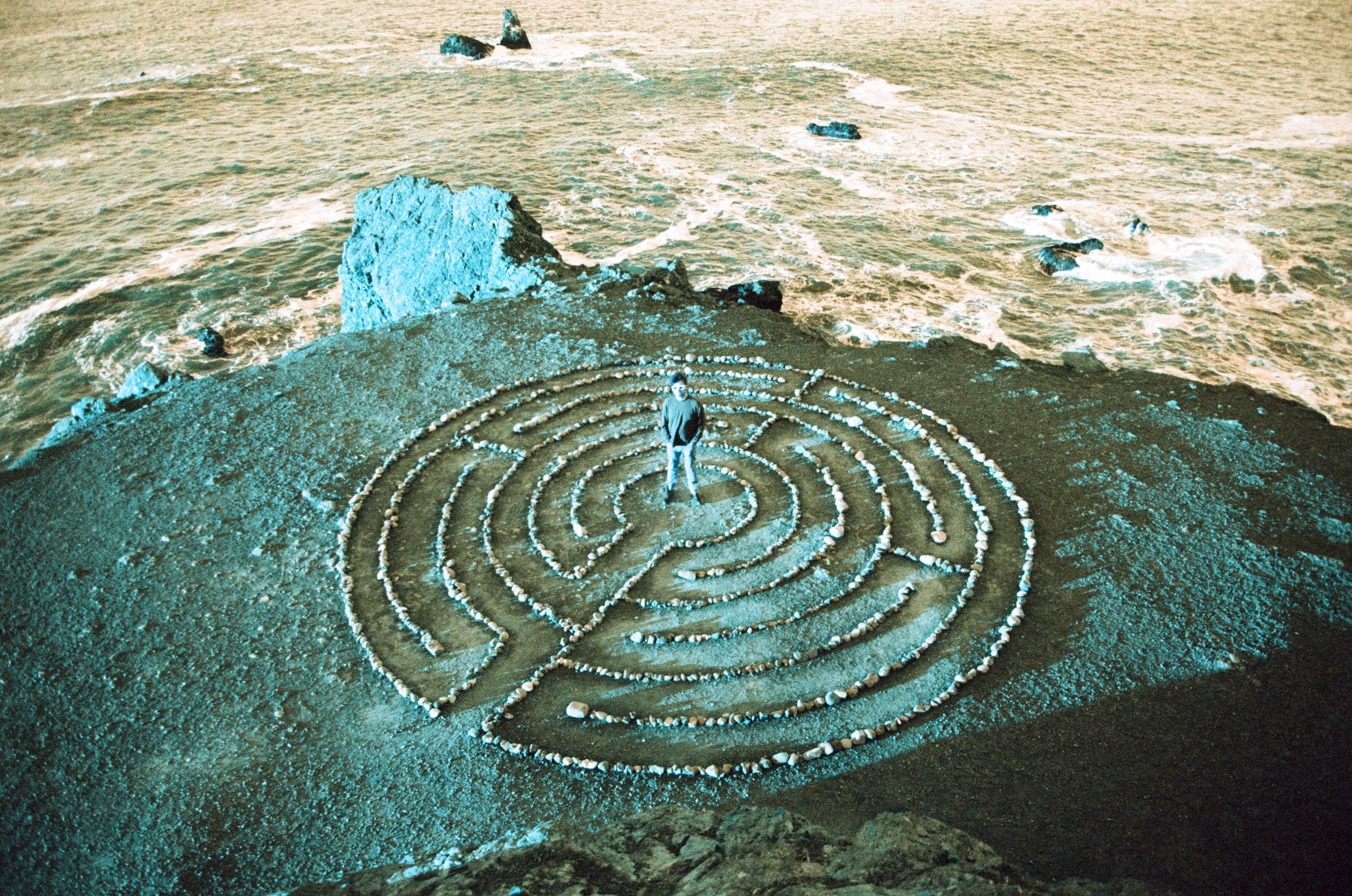 A solitary figure standing inside a stone labyrinth on coastal bluffs above the ocean